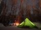A man next to a fat bike in the woods leans over a campfire and tent at night with the stars out.