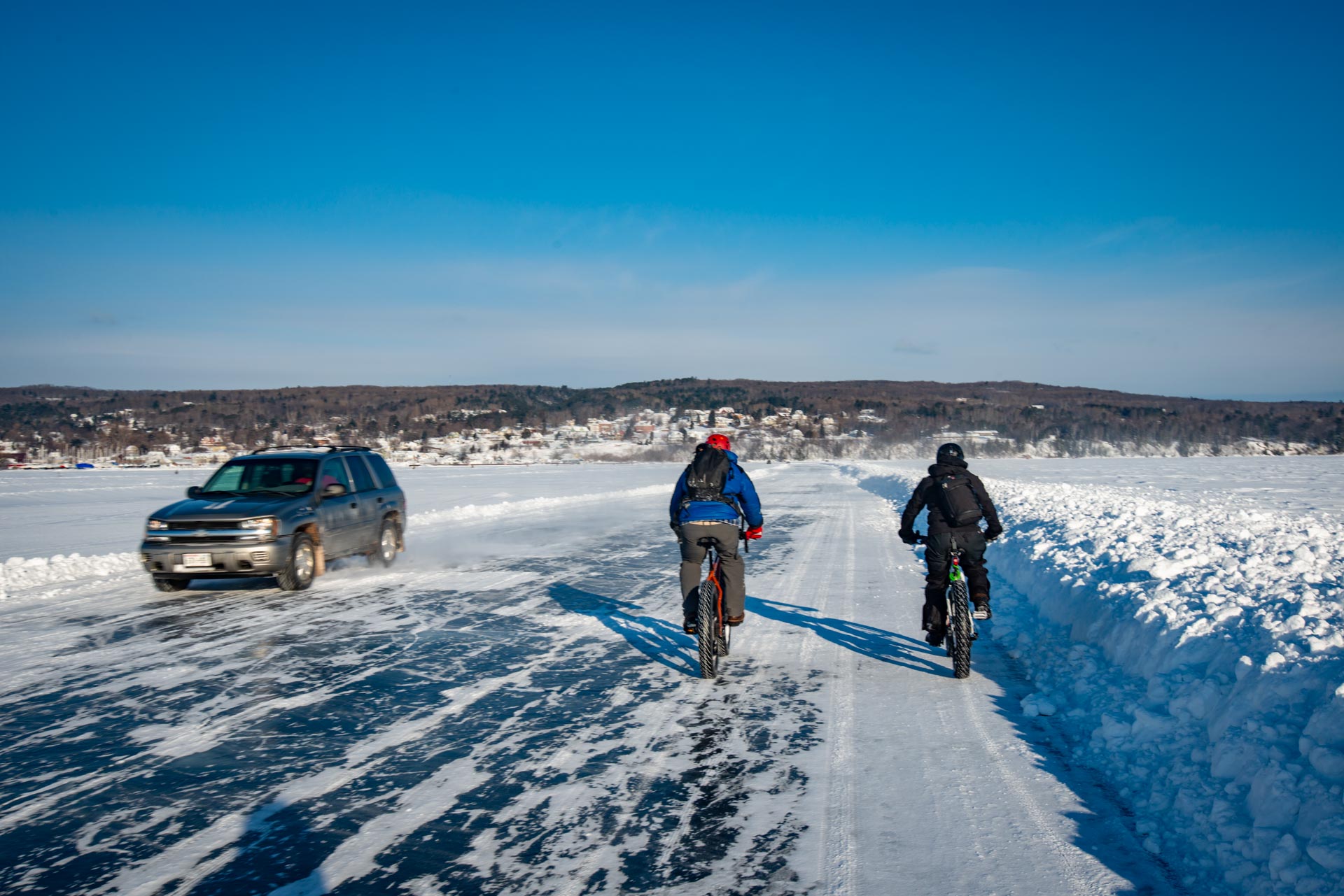Two people riding bikes past an SUV on the ice road to Madeline Island